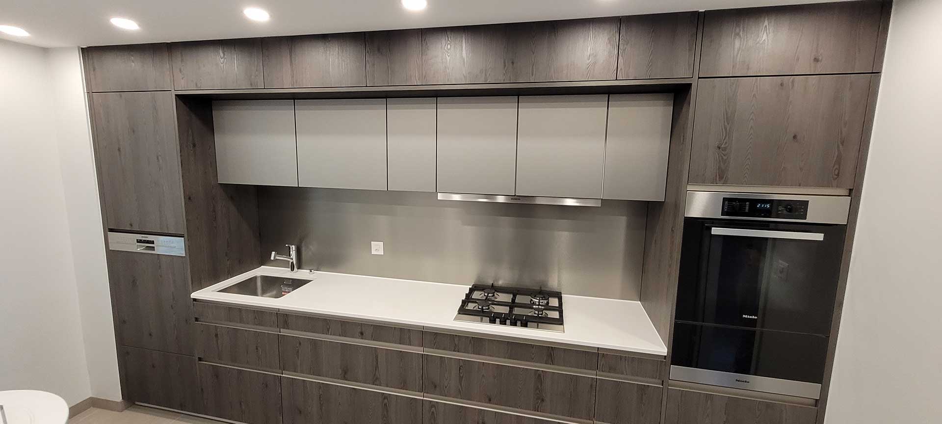 Cuisine linéaire en façades bois foncé, niche claire, crédence en inox brossé, plan de travail blanc et four Miele encastré