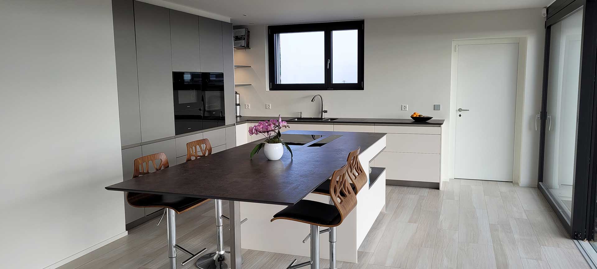 Cuisine ouverte avec colonnes anthracite, îlot-table au plateau sombre, tabourets de bar et grande baie vitrée